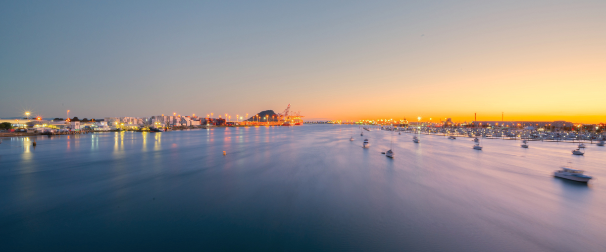 The Gold House® — Tauranga Harbour at sunset, New Zealand Panoramic view of Tauranga Harbour at sunset with Mount Maunganui, port cranes, and marina lights — The Gold House® precious metals brokerage, New Zealand